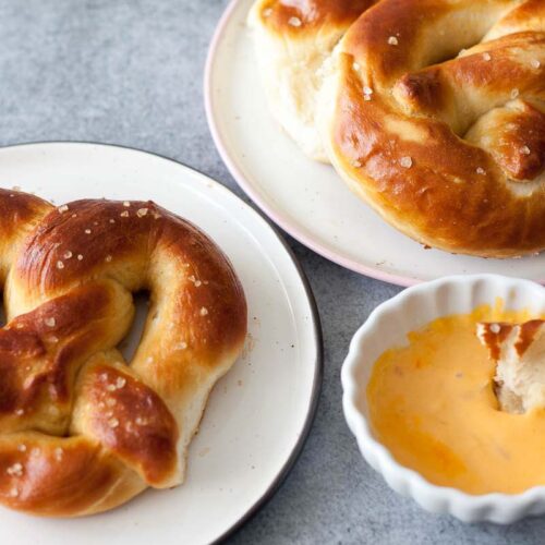 Two golden-brown soft pretzels on white plates, sprinkled with coarse salt, served alongside a small dish of creamy cheese dip with a piece of pretzel dipped inside.