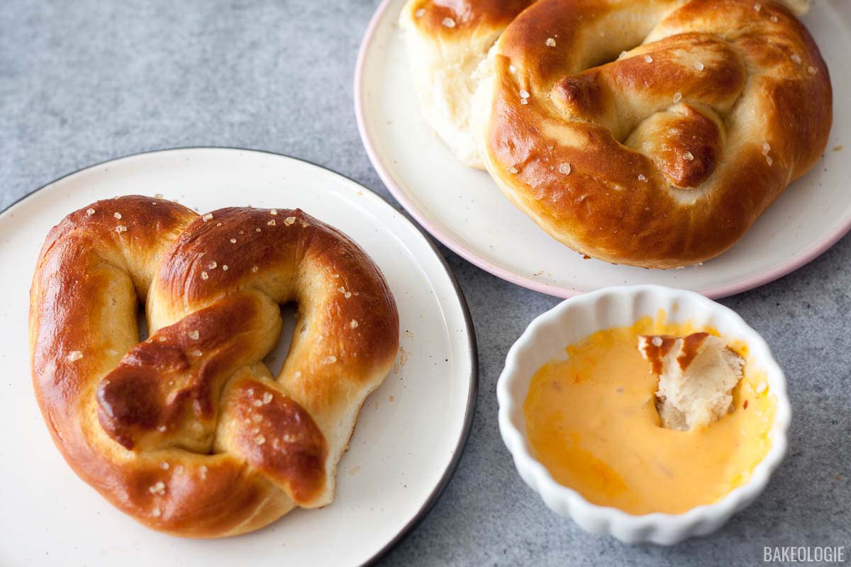 Two golden-brown soft pretzels on white plates, sprinkled with coarse salt, served alongside a small dish of creamy cheese dip with a piece of pretzel dipped inside.