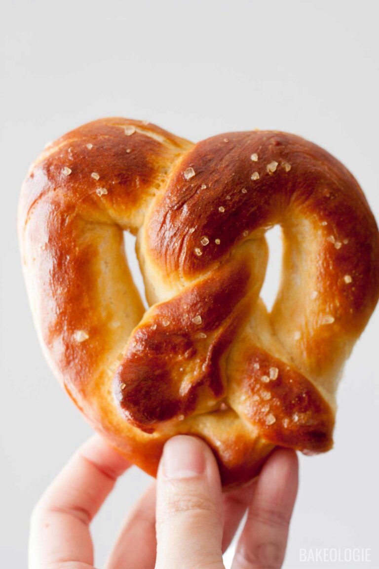 Close-up of a freshly baked soft pretzel with a golden-brown crust, sprinkled with coarse salt, held by a hand against a light background.