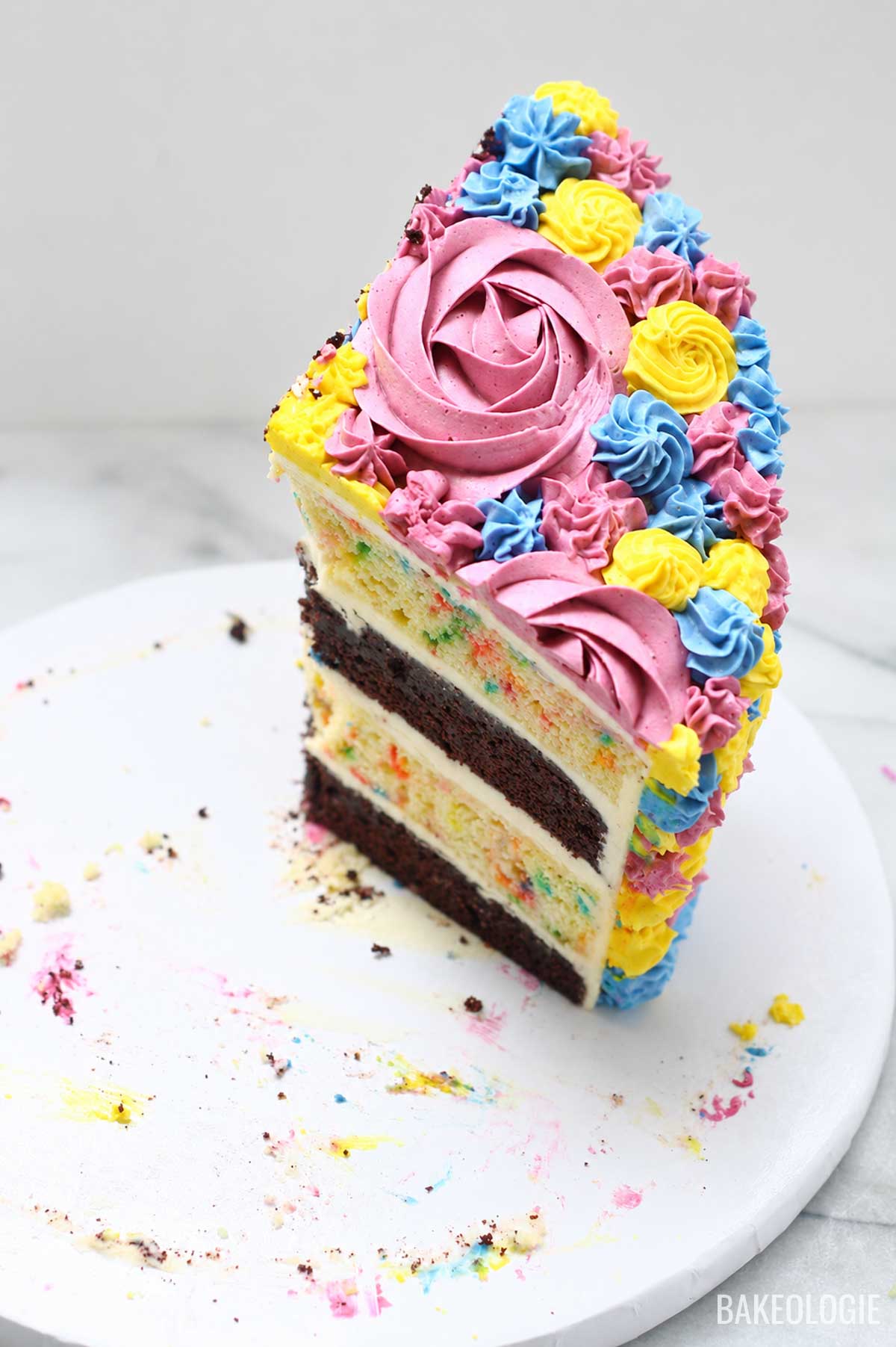 Slice of layered chocolate and funfetti cake decorated with pink, blue, and yellow buttercream rosettes on a white cake stand.