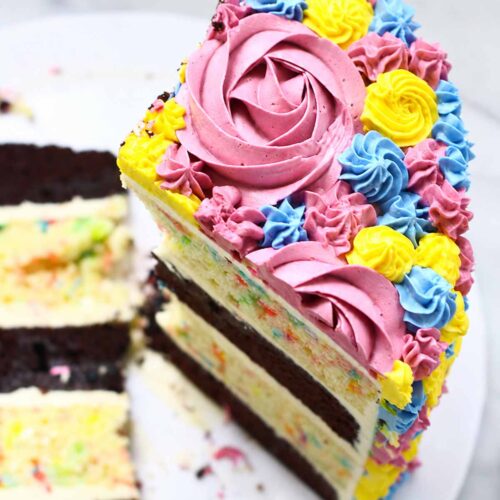 Close-up of colorful buttercream cake slice with pink rosettes, blue swirls, and yellow flowers on a marble background.