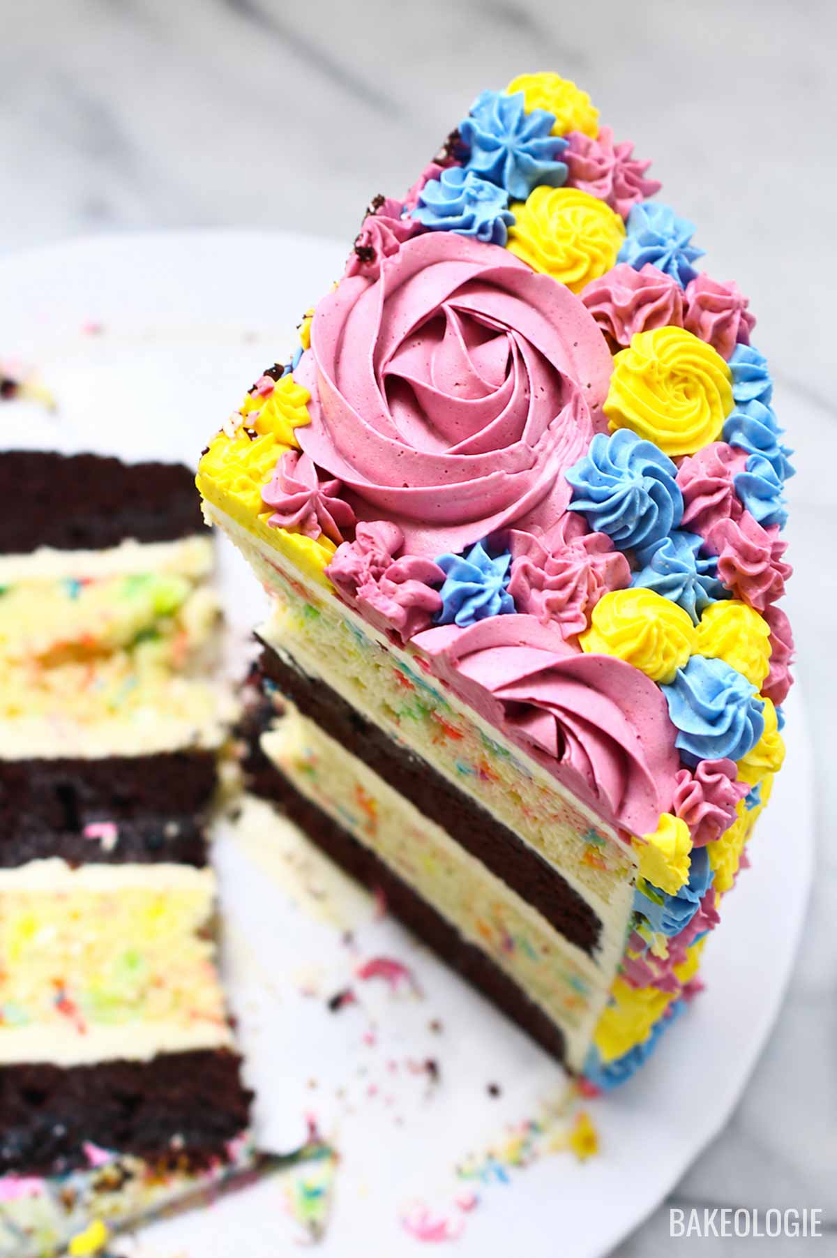 Close-up of colorful buttercream cake slice with pink rosettes, blue swirls, and yellow flowers on a marble background.