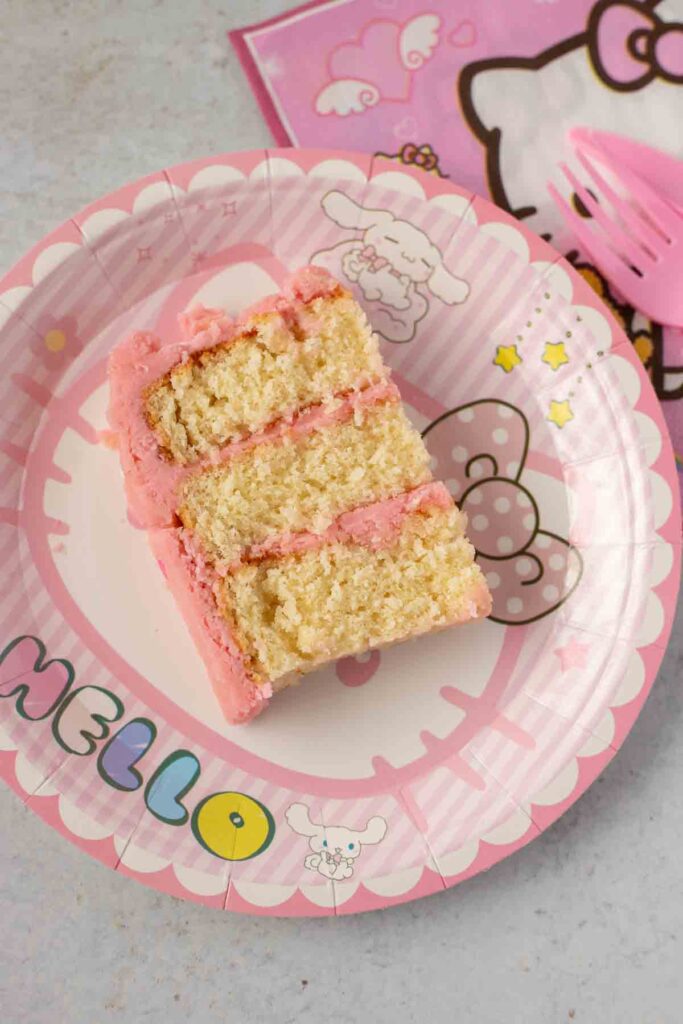a slice of vanilla cake with pink buttercream on a hello kitty plate with hello kitty napkin on the side and pink fork