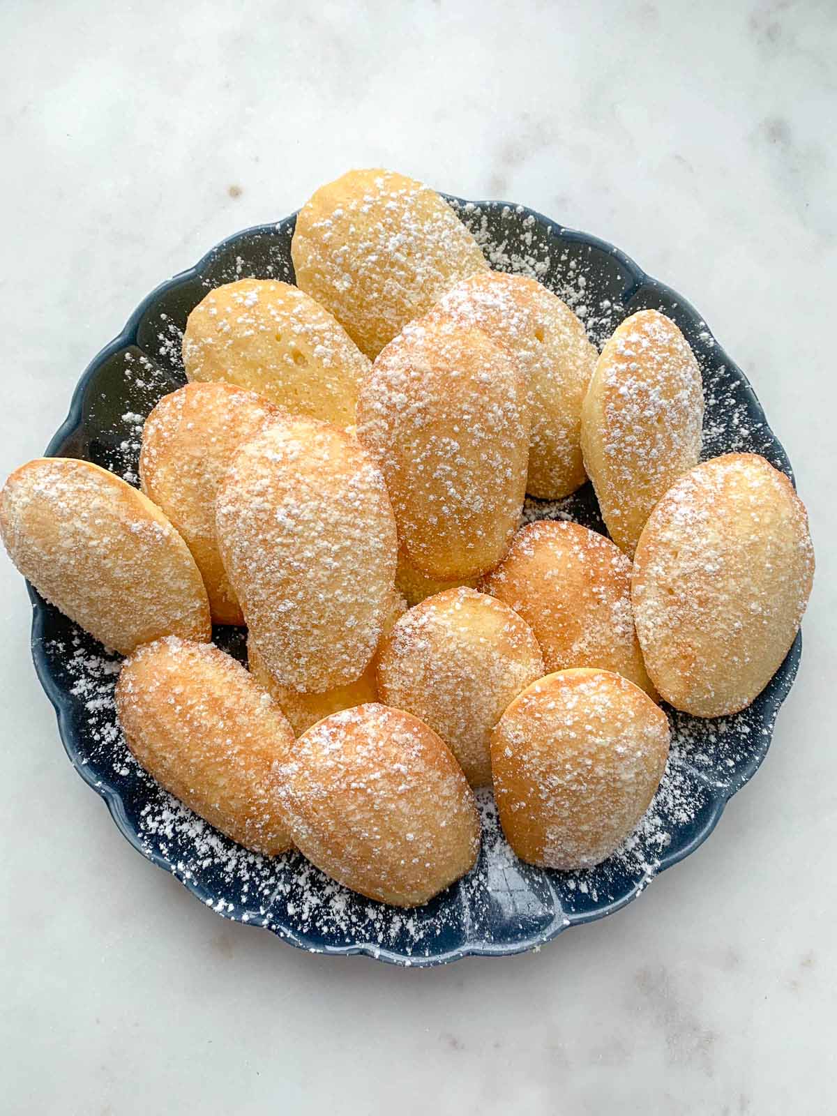 Classic French madeleines with golden shells and signature humps, lightly dusted with powdered sugar and arranged in a fluted serving dish.