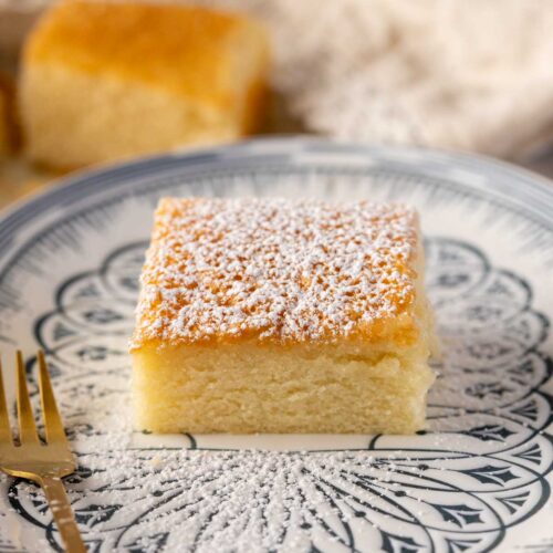 A slice of Hot Milk Cake dusted with powdered sugar, served on a decorative plate. The cake has a golden top and a soft, fluffy texture, accompanied by a gold fork.