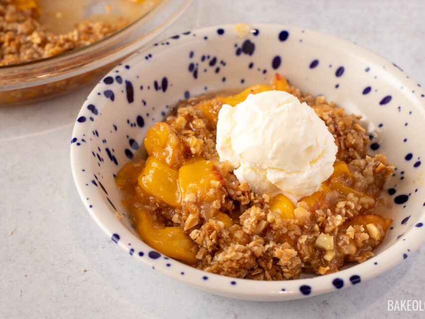 Bowl of homemade peach crisp topped with a scoop of vanilla ice cream, featuring golden, juicy peaches and a crunchy oat topping, served in a white bowl with blue speckles.