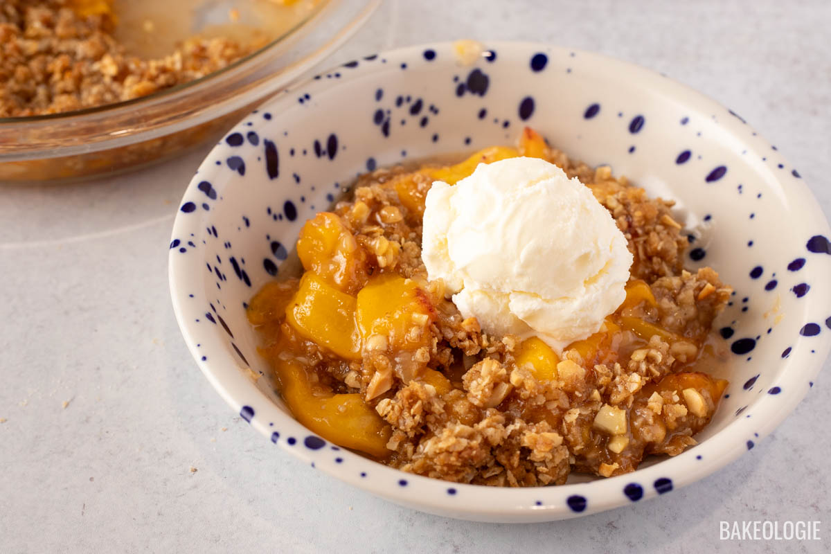 Bowl of homemade peach crisp topped with a scoop of vanilla ice cream, featuring golden, juicy peaches and a crunchy oat topping, served in a white bowl with blue speckles.