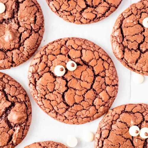 A batch of fudgy brownie cookies with crackly tops, scattered across a white background. Each cookie is decorated with two small chocolate eyes, giving them a fun, whimsical look. Some cookies are whole, while one cookie is broken in half, showing a soft and rich interior. White chocolate chips are scattered around the cookies for added texture.