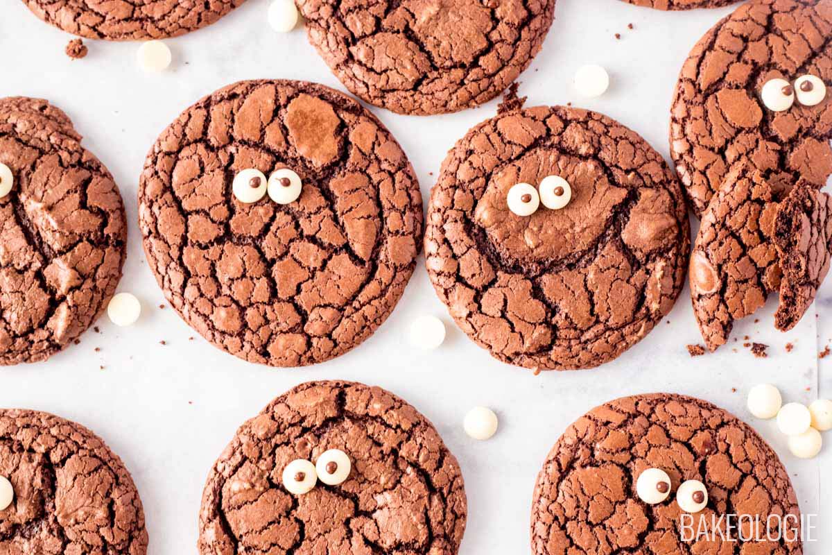 A batch of fudgy brownie cookies with crackly tops, scattered across a white background. Each cookie is decorated with two small candy eyes, giving them a fun, whimsical look. Some cookies are whole, while one cookie is broken in half, showing a soft and rich interior. White chocolate chips are scattered around the cookies for added texture.