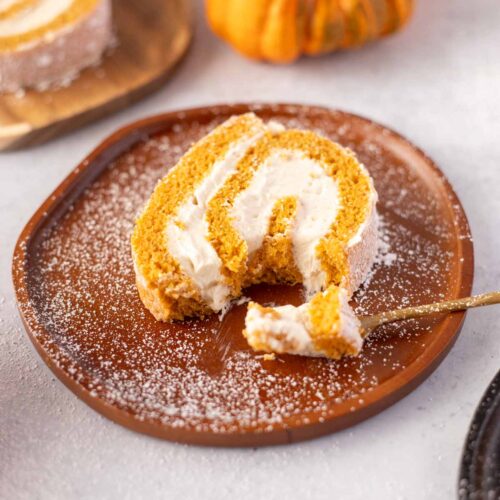 A close-up of a slice of pumpkin roll with cream cheese frosting on a brown plate, dusted with powdered sugar. A forkful of the cake has been taken out, revealing the creamy filling. In the background, there is another slice of pumpkin roll on a wooden plate, along with small decorative pumpkins, enhancing the fall theme.