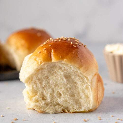 Close-up of a single Japanese milk bread roll with a soft, fluffy interior, topped with sesame seeds, showing its tender crumb texture on a light background.
