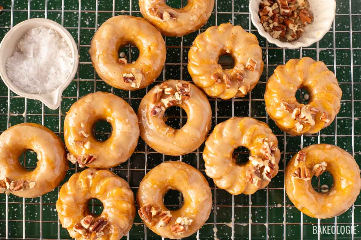 A batch of round, golden-brown pumpkin donuts with a salted maple glaze, topped with chopped pecans, arranged on a wire cooling rack. To the left is a small white dish filled with flaky sea salt, and to the top right, a small bowl containing more chopped pecans. The donuts are glossy from the glaze, creating an inviting and delicious look.