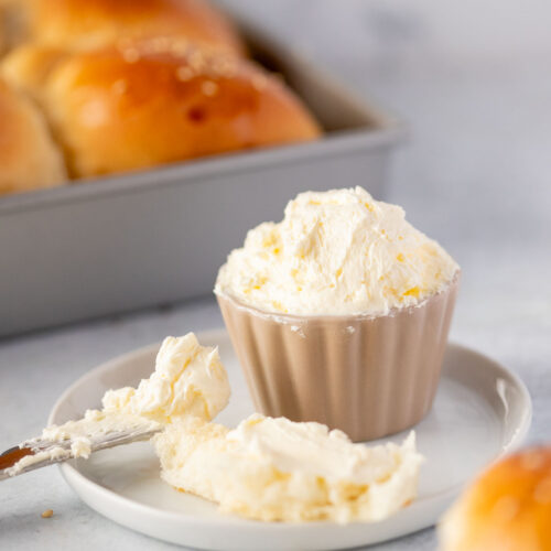 A small dish of fluffy whipped butter sits on a white plate with a knife and a piece of bread spread with butter. In the background, a tray of freshly baked golden bread rolls is slightly blurred. The scene emphasizes the soft, spreadable texture of the whipped butter, perfect for warm bread.