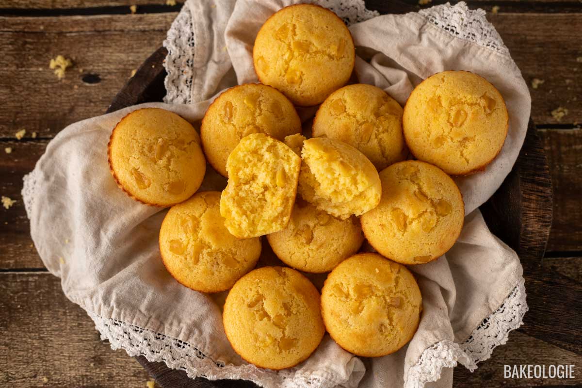 Basket of golden cornbread muffins, with one muffin split open to reveal a moist, fluffy interior, resting on a linen cloth with lace trim on a rustic wooden background.
