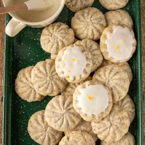 A green tray filled with lemon poppy seed cookies, some topped with a lemon glaze and zest. A small bowl of glaze with a pastry brush rests in the corner of the tray.
