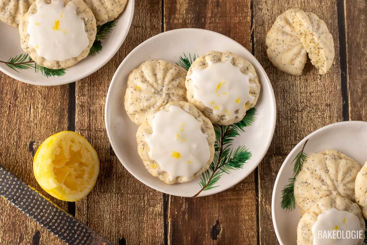 Lemon poppy seed cookies with a glaze, served on festive plates with pine designs, placed on a rustic wooden surface. A zested lemon and a grater are visible beside the cookies.