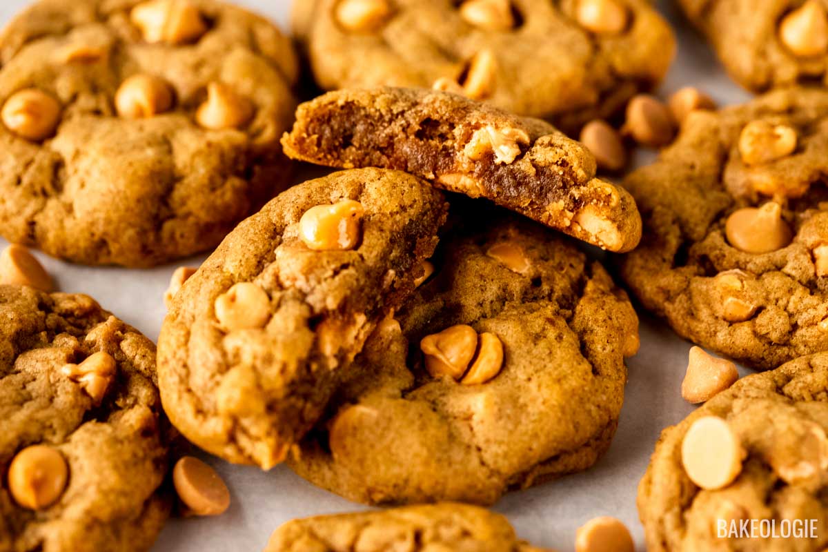 A pile of pumpkin butterscotch cookies with one cookie broken in half to showcase its chewy texture and generous butterscotch chip filling.