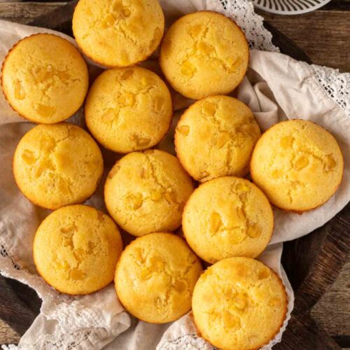 Top view of golden cornbread muffins arranged on a linen-lined basket, accompanied by a small dish of butter and a bowl of corn kernels, set on a rustic wooden table.