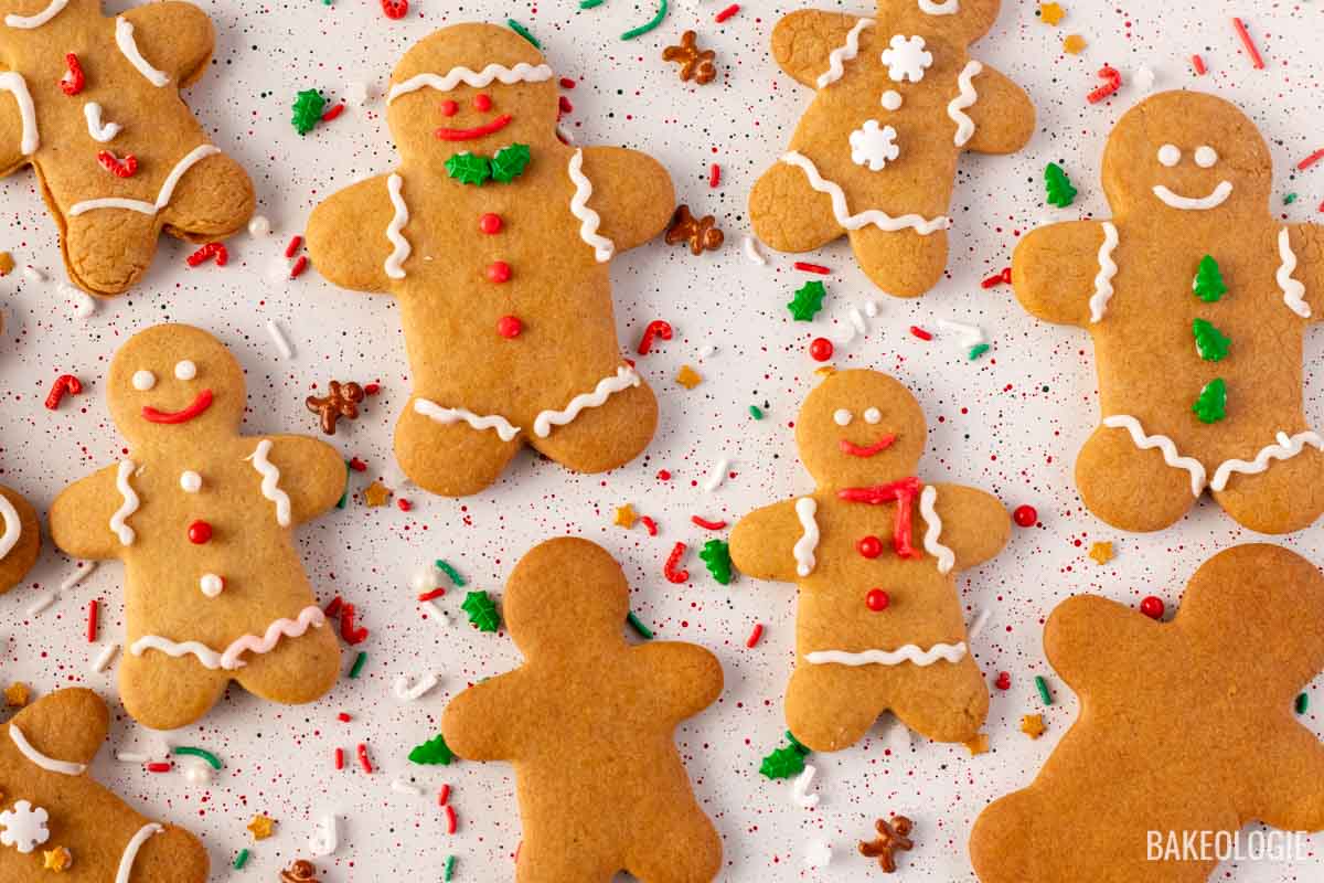 Festive gingerbread cookies shaped like little people, decorated with white icing, red buttons, and colorful holiday sprinkles on a white surface sprinkled with red and green confetti.