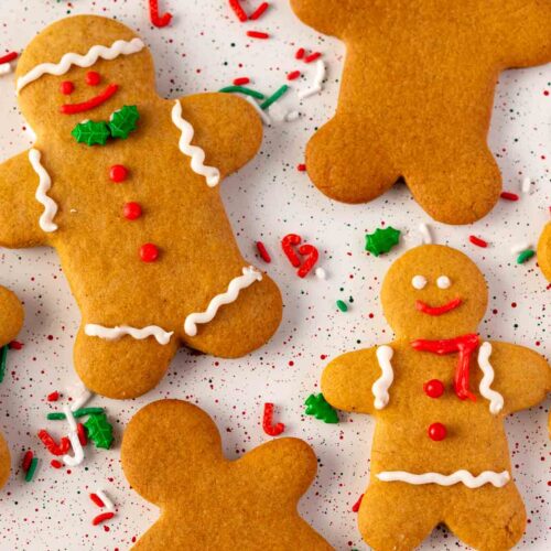 Gingerbread cookies shaped like people, some decorated with white icing, red buttons, and festive holiday sprinkles, displayed on a white surface with red, green, and white confetti.