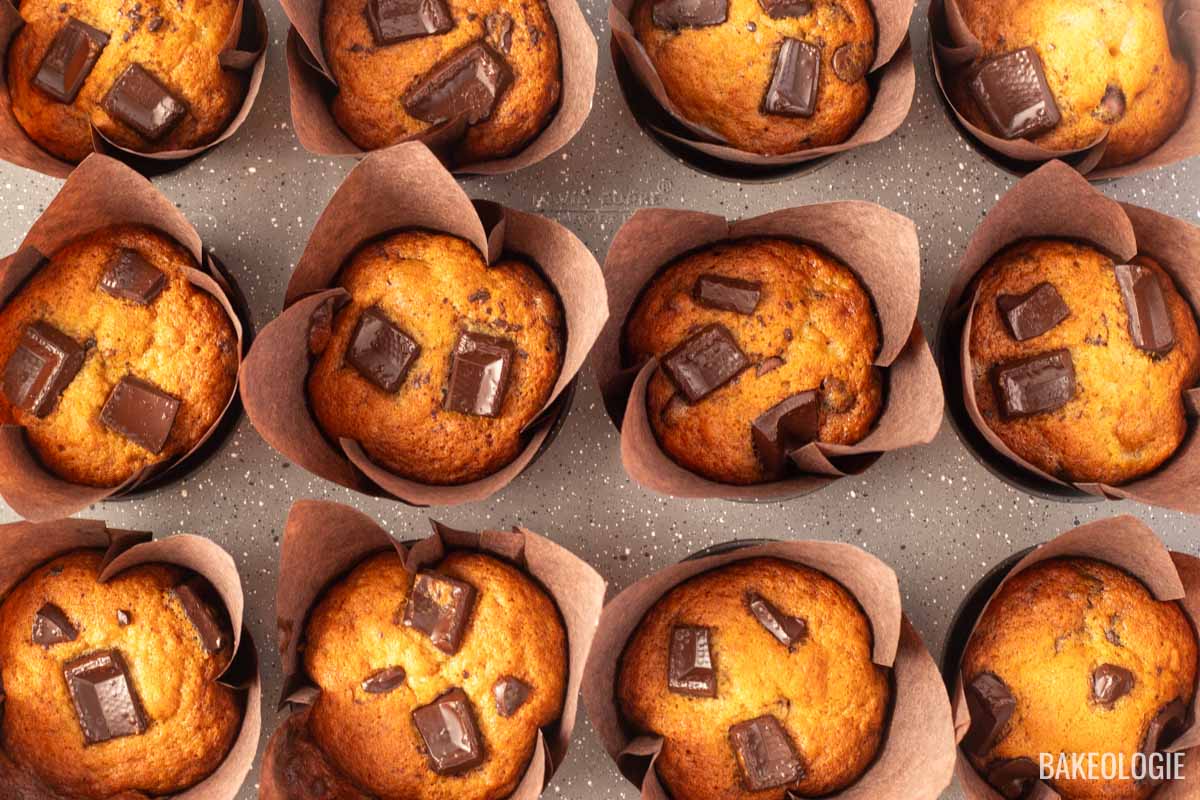 A batch of freshly baked chocolate chunk banana muffins in tulip-style liners. The muffins are golden brown with large, melted chocolate chunks on top, showcasing their moist texture and rich chocolatey goodness.