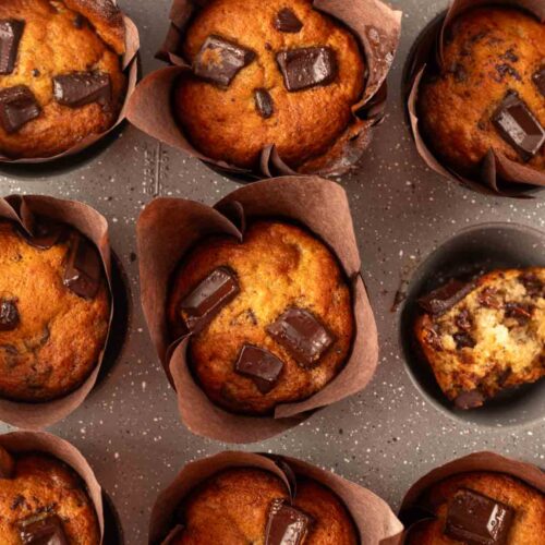 A batch of freshly baked chocolate chunk banana muffins in tulip-style liners, some whole, some halved. The muffins are golden brown with large, melted chocolate chunks on top, showcasing their moist texture and rich chocolatey goodness.