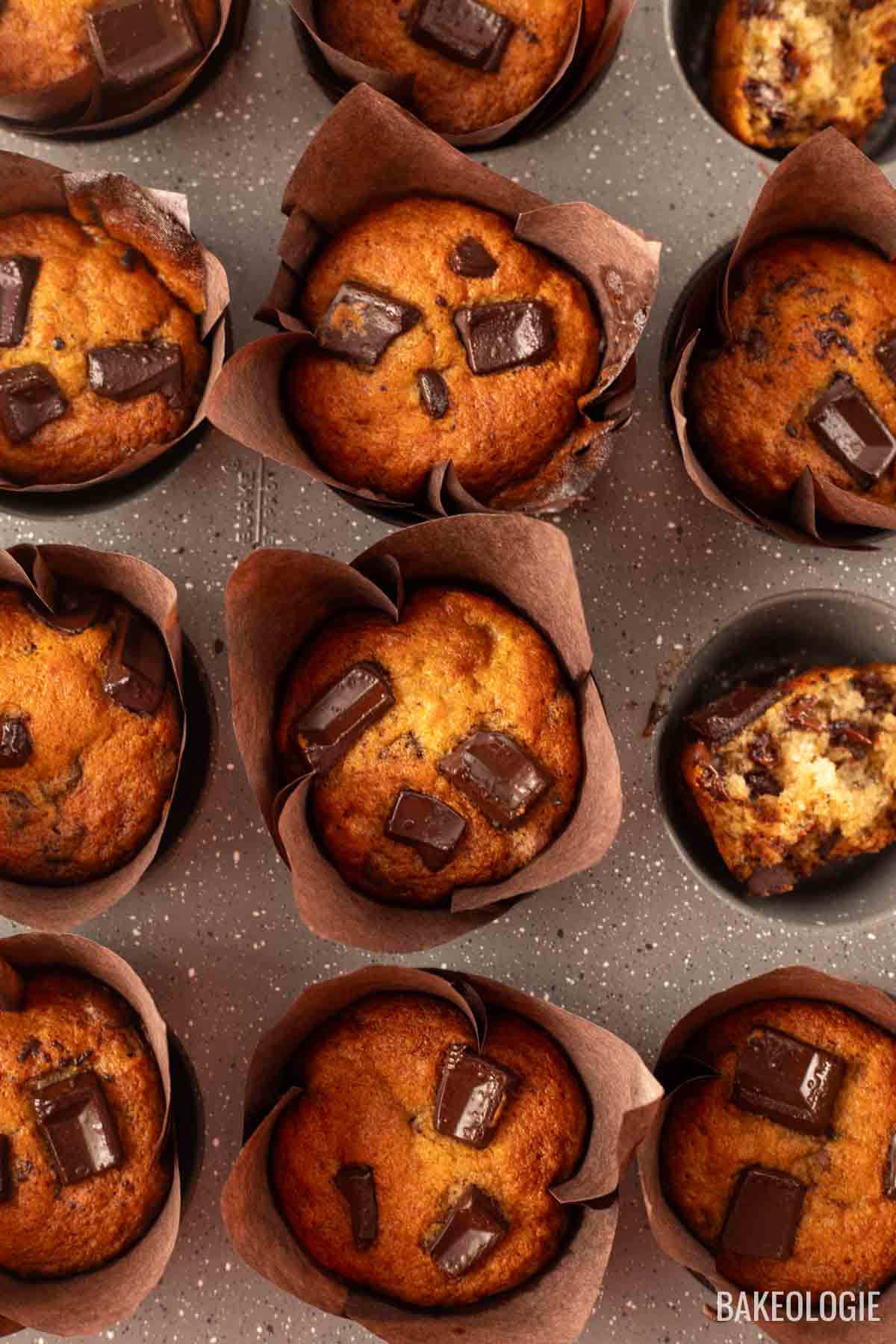 A batch of freshly baked chocolate chunk banana muffins in tulip-style liners, some whole, some halved. The muffins are golden brown with large, melted chocolate chunks on top, showcasing their moist texture and rich chocolatey goodness.