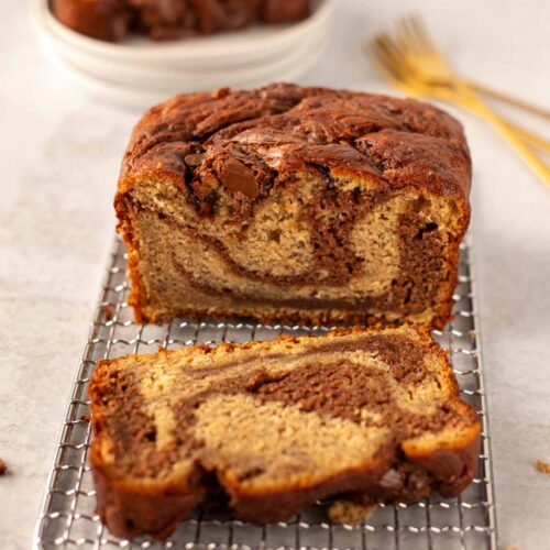 A loaf of marbled banana bread with chocolate swirls sits on a cooling rack, with one slice cut to reveal its soft and rich texture. In the background, stacked white plates and gold forks add a touch of elegance to the scene.