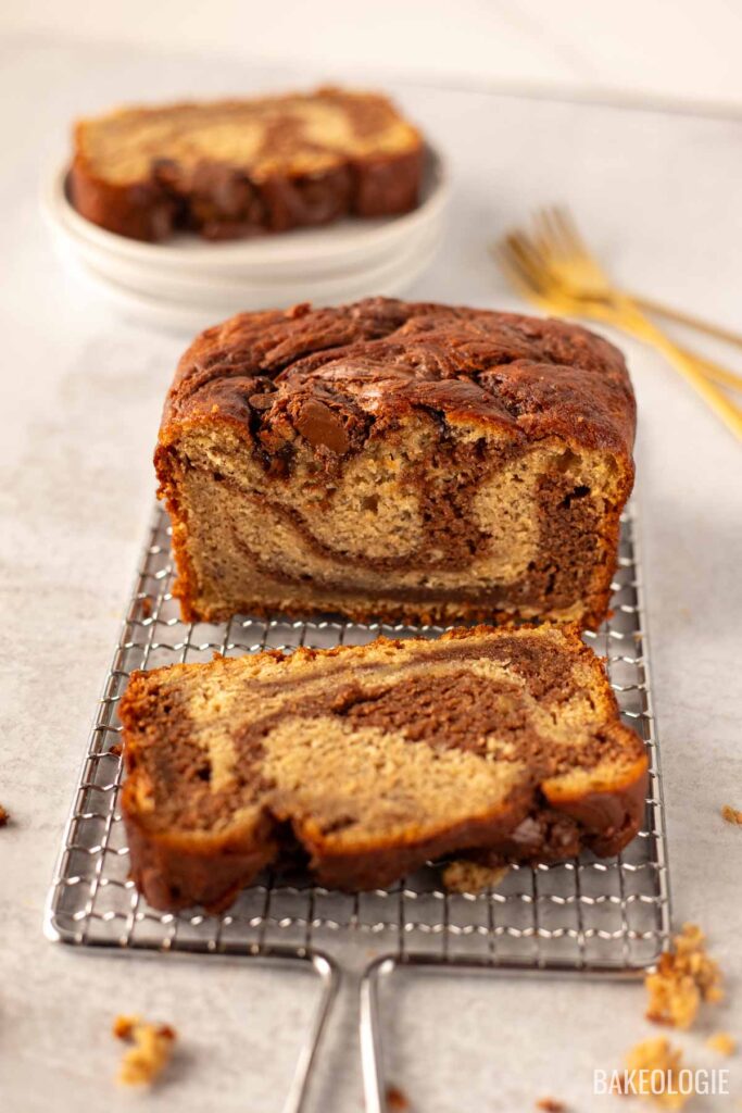 A loaf of marbled banana bread with chocolate swirls sits on a cooling rack, with one slice cut to reveal its soft and rich texture. In the background, stacked white plates and gold forks add a touch of elegance to the scene.