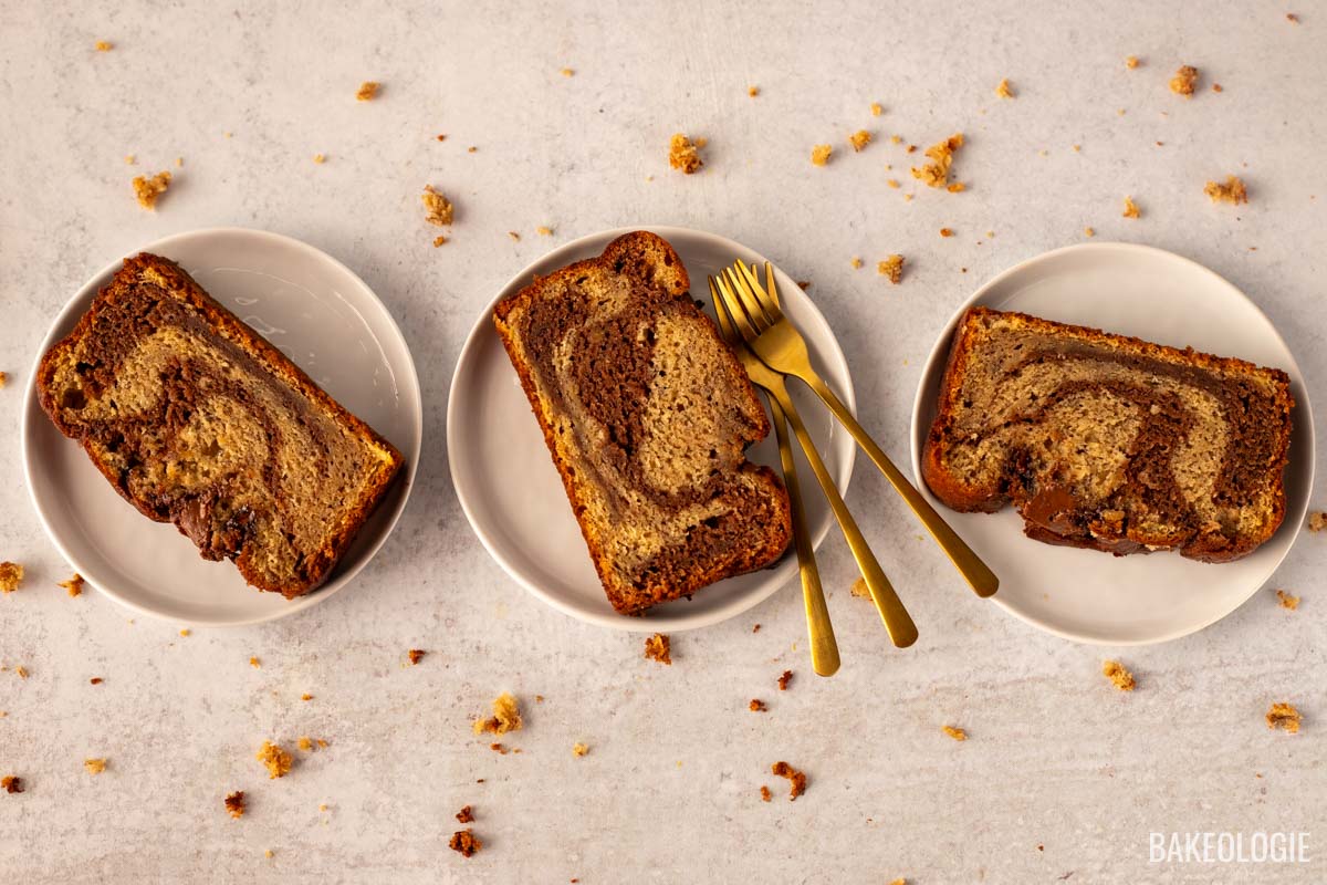 Three slices of marbled banana bread with chocolate swirls served on white plates, accompanied by gold forks on a light marble surface. Crumbs are scattered around for a rustic touch.