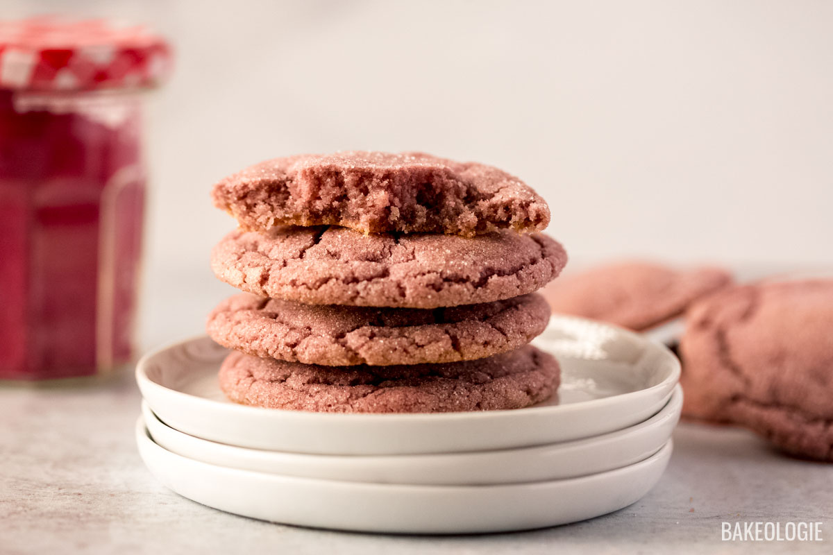 A stack of four pink sugar cookies on a set of white plates, with the top cookie having a bite taken out. The cookies have a slightly cracked surface and are coated in sugar. In the blurred background, there is a jar of freeze dried strawberries with a checkered lid and additional cookies resting on a countertop. The lighting is soft and warm, highlighting the texture of the cookies.