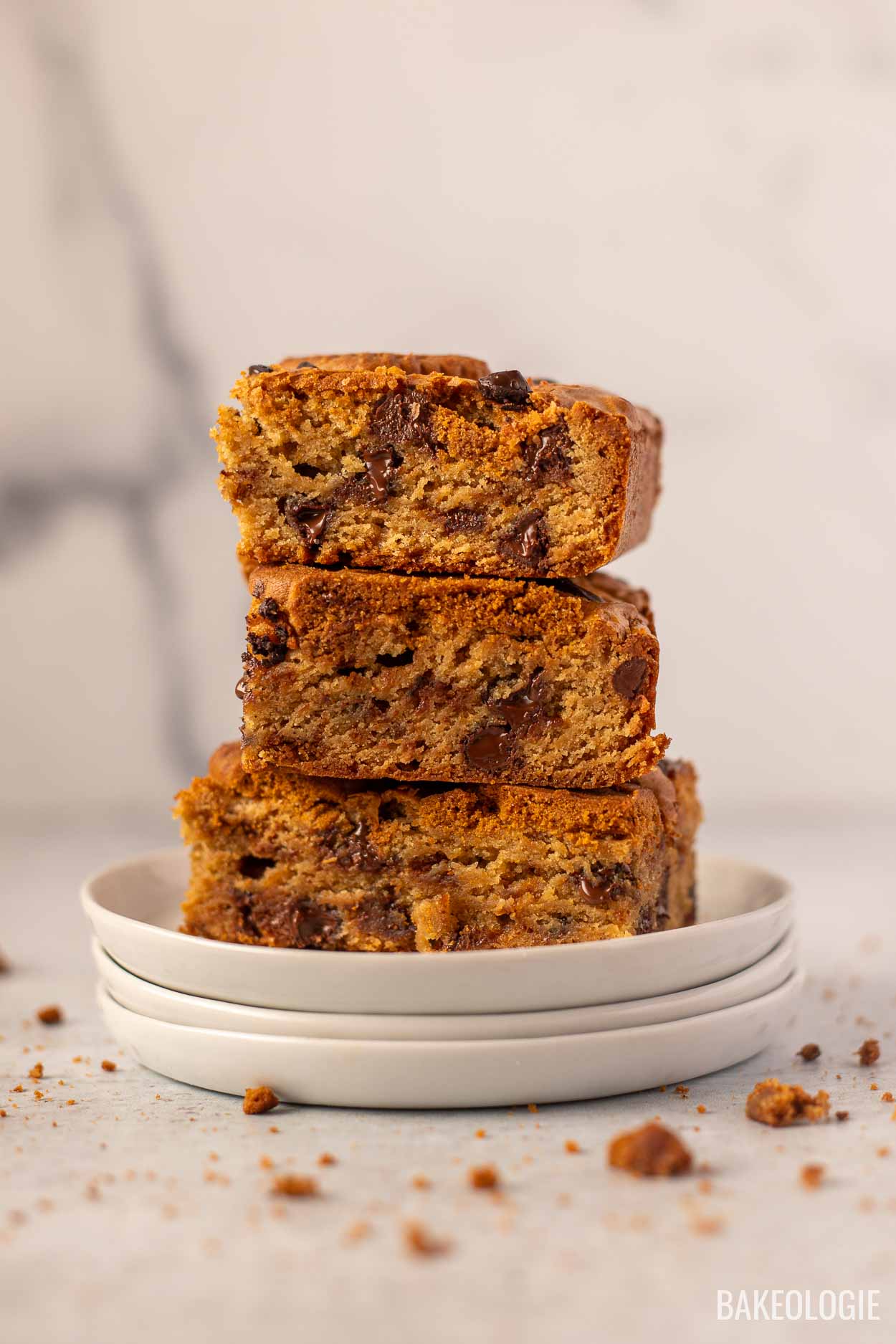 A stack of three thick Biscoff blondie bars on a small white plate, showing their soft, gooey texture with melted chocolate chunks throughout. The background is blurred, with a few crumbs scattered on the surface.