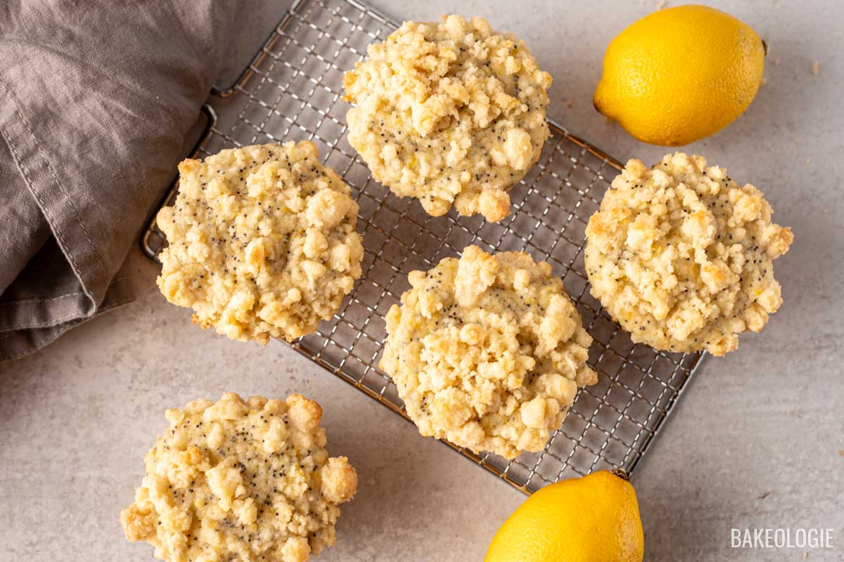 Five lemon poppy seed muffins with a crumbly streusel topping on a cooling rack, accompanied by fresh lemons on a light, textured surface. A gray linen napkin is partially visible on the left side.