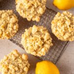 Five lemon poppy seed muffins with a crumbly streusel topping on a cooling rack, accompanied by fresh lemons on a light, textured surface. A gray linen napkin is partially visible on the left side.
