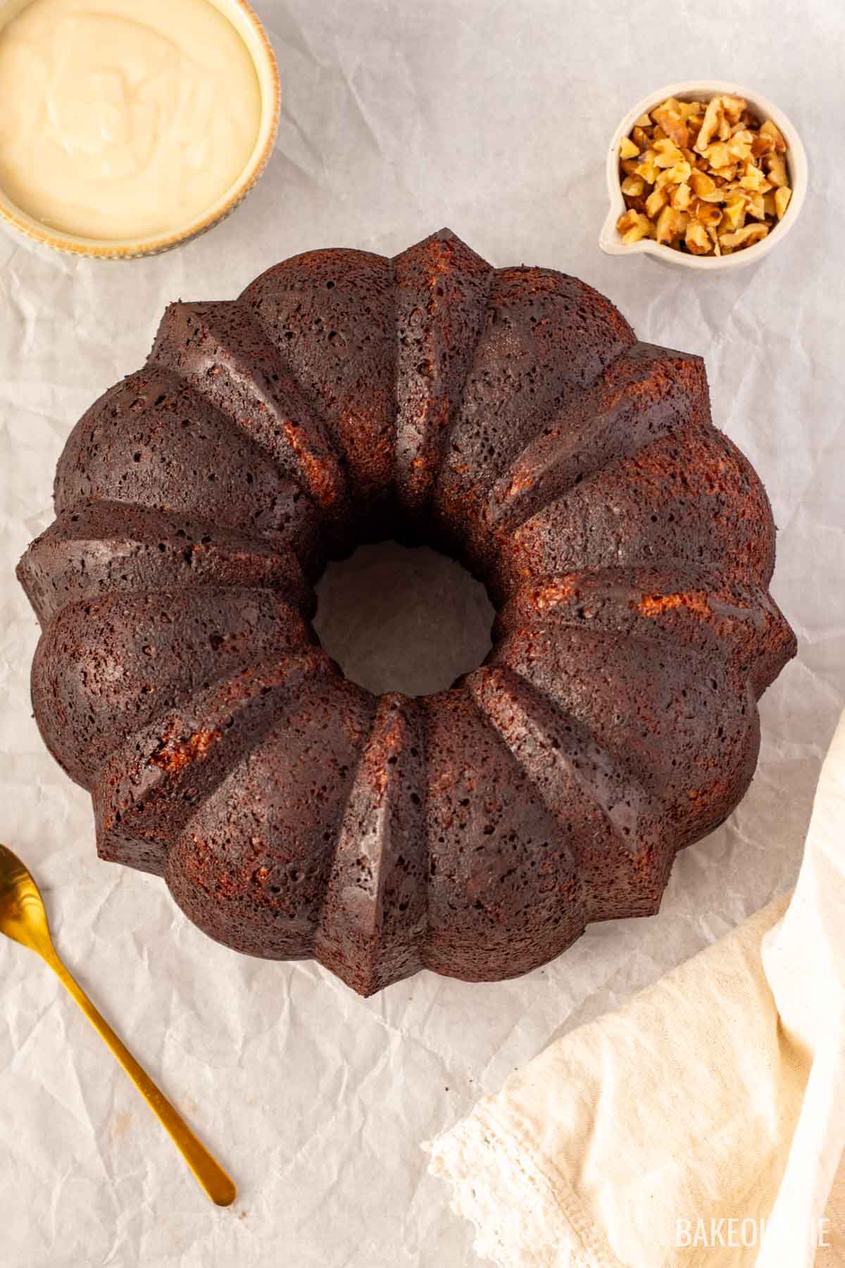 Overhead view of an unglazed carrot Bundt cake sitting on parchment paper, with a small bowl of chopped walnuts and a golden spoon beside it.