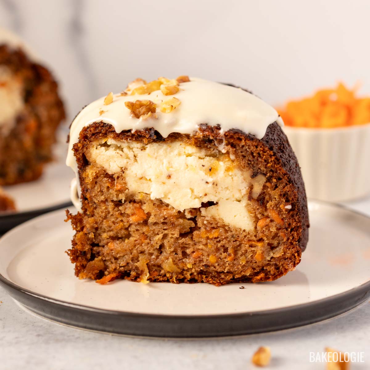 A close-up of a slice of cheesecake stuffed carrot Bundt cake showing the creamy cheesecake filling inside, topped with cream cheese glaze and chopped walnuts, served on a white plate with a side of shredded carrots.