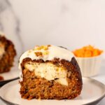 A close-up of a slice of carrot Bundt cake revealing the creamy cheesecake filling inside, topped with a cream cheese glaze and sprinkled with chopped walnuts.