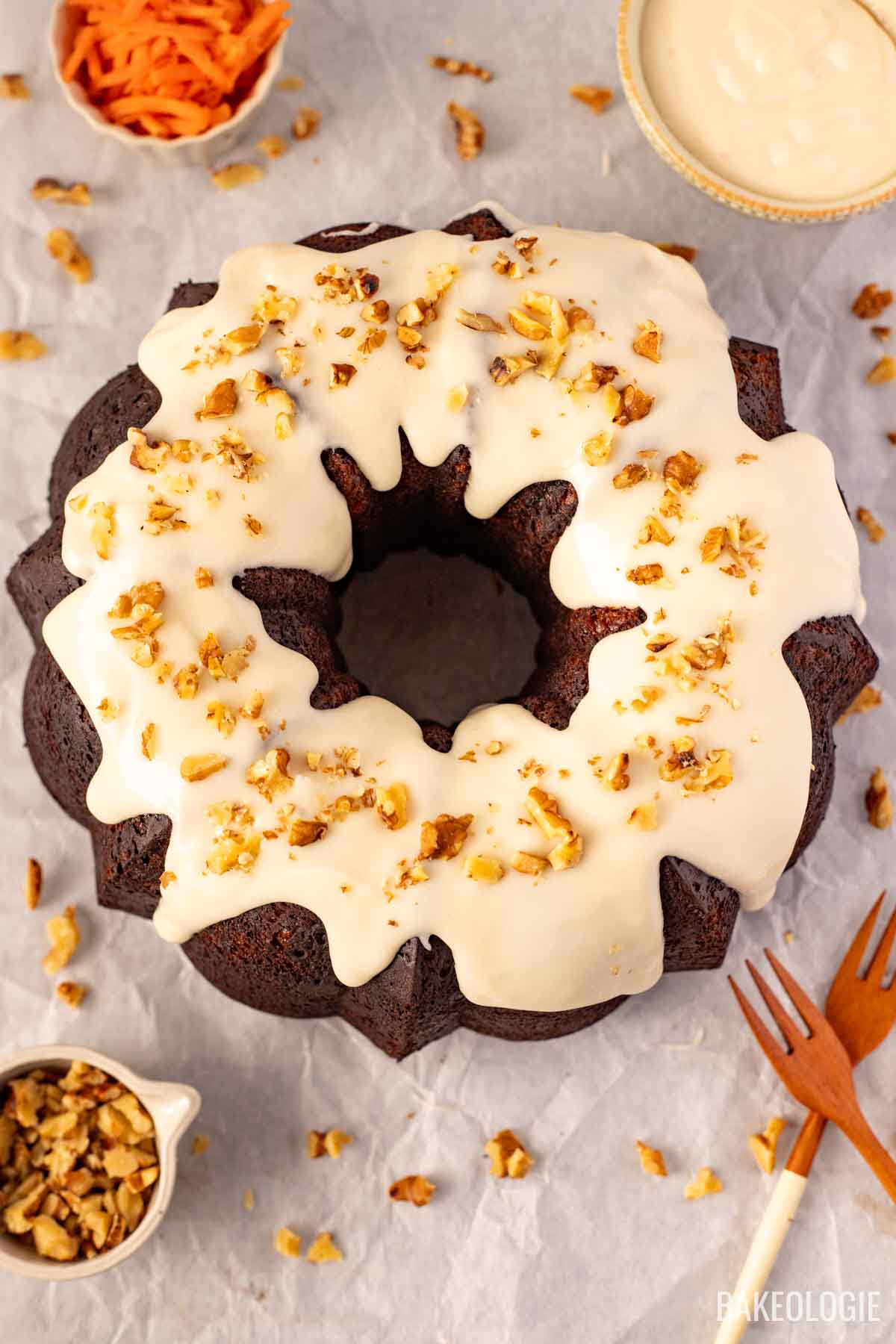 Overhead shot of a cheesecake stuffed carrot Bundt cake drizzled with cream cheese glaze and topped with chopped walnuts, surrounded by shredded carrots and a wooden fork.