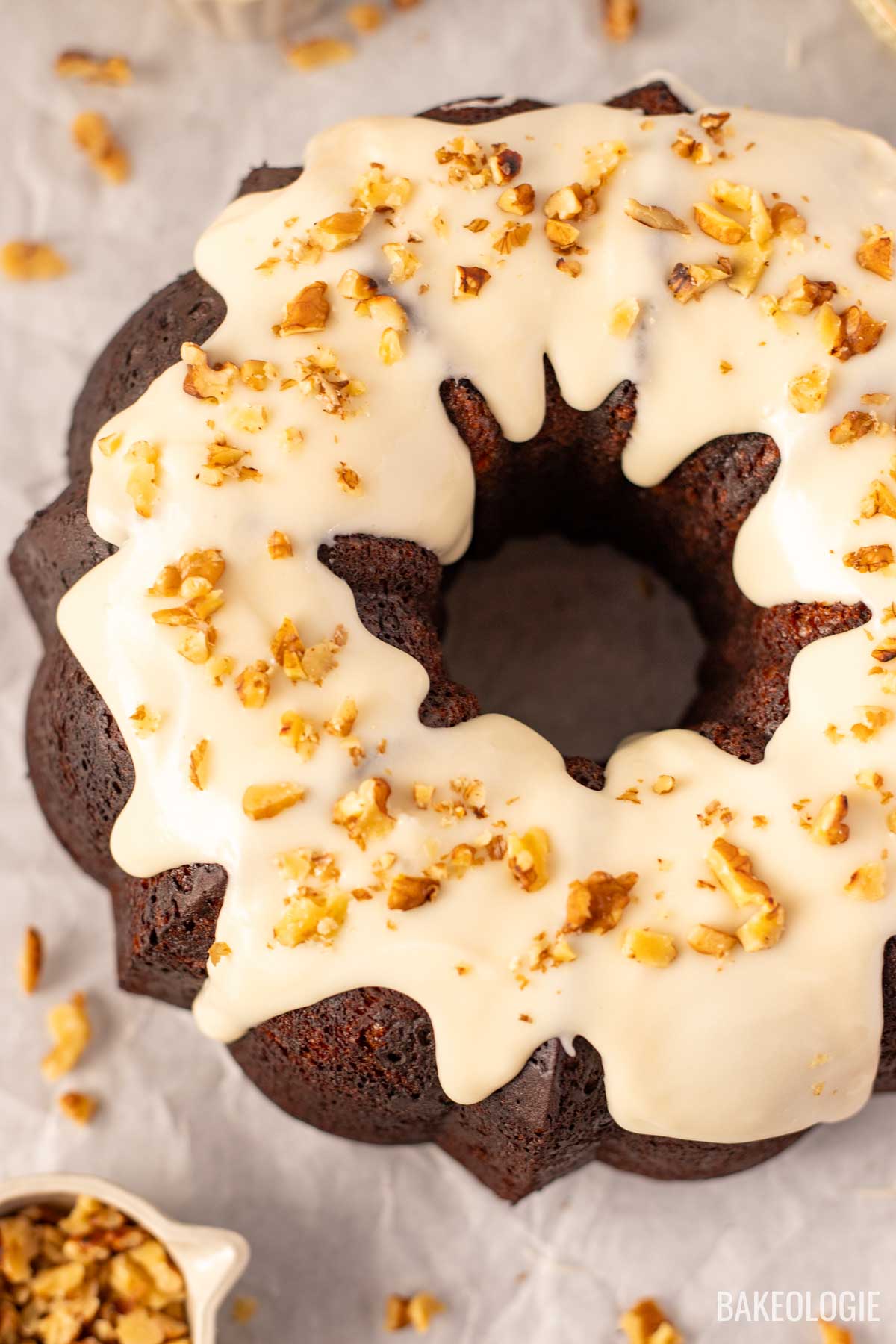Overhead view of a cheesecake-stuffed carrot Bundt cake drizzled with cream cheese glaze and topped with chopped walnuts, resting on parchment paper with scattered walnuts around it.