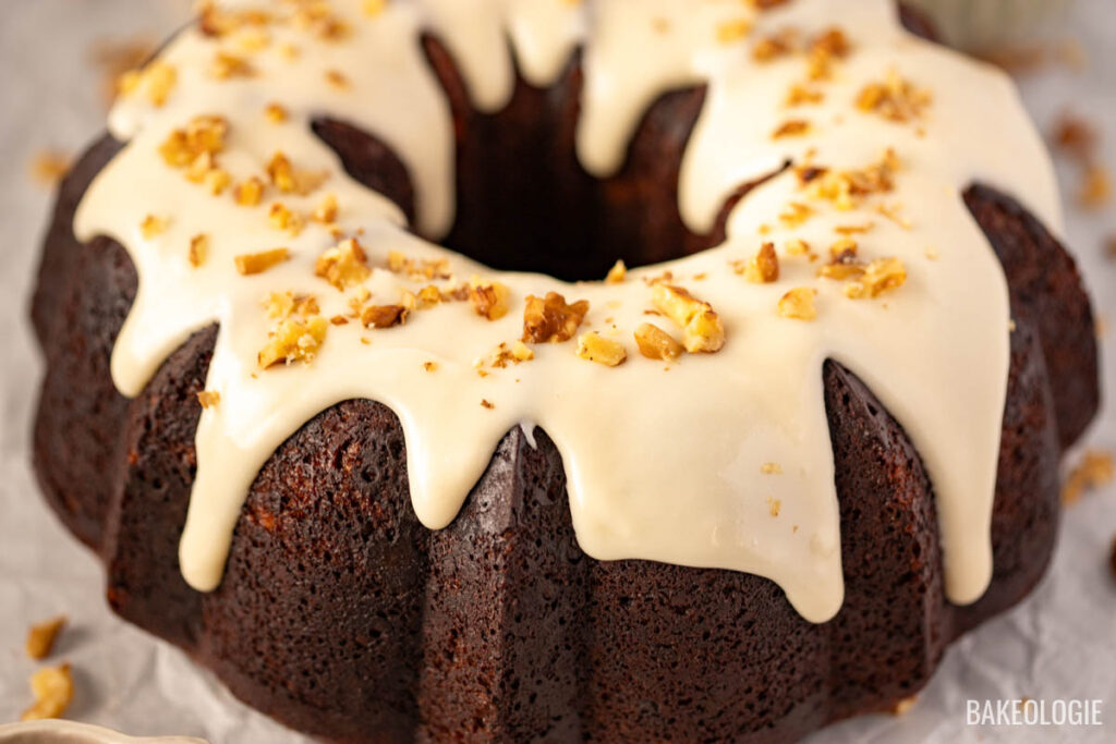 Close-up view of a cheesecake stuffed carrot Bundt cake drizzled with cream cheese glaze and topped with chopped walnuts, resting on parchment paper.