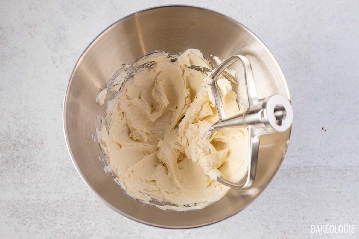 Close-up of a stand mixer with a paddle attachment, mixing a creamy cream cheese glaze in a stainless steel bowl.