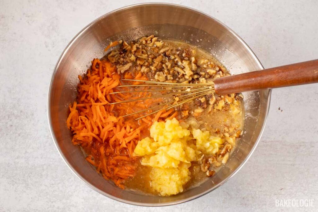 Close-up of a bowl with shredded carrots, chopped walnuts, and pineapple being mixed with wet ingredients using a whisk.