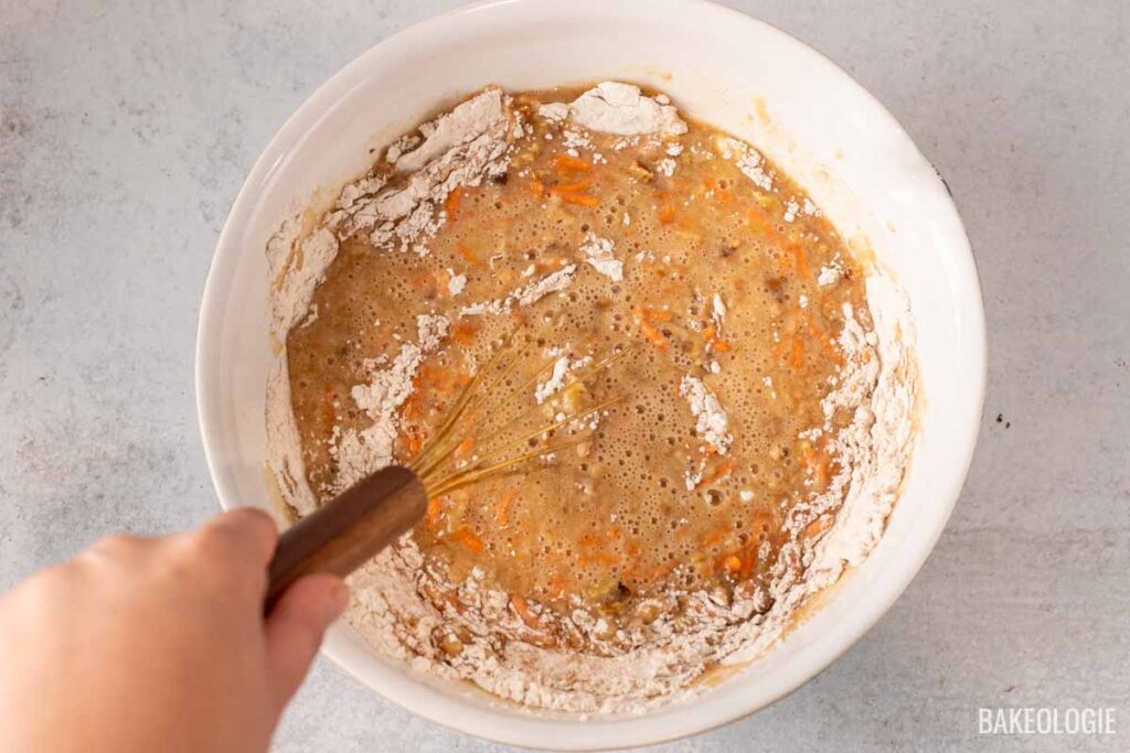 A hand using a whisk to mix the wet ingredients and shredded carrots into the dry ingredients in a large bowl, creating the carrot cake batter.