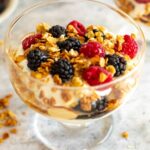 Close-up side view of a Greek yogurt parfait served in a clear glass, highlighting layers of creamy yogurt, golden granola clusters, and vibrant fresh blackberries and raspberries.