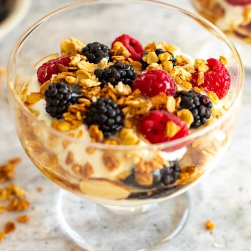 Close-up side view of a Greek yogurt parfait served in a clear glass, highlighting layers of creamy yogurt, golden granola clusters, and vibrant fresh blackberries and raspberries.