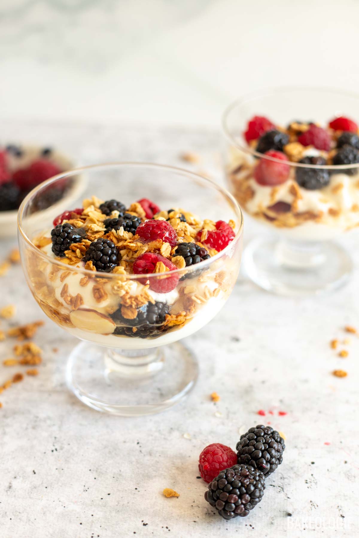 Front view of a glass cup filled with layers of Greek yogurt, granola, blackberries, and raspberries, with a few berries and scattered granola pieces on the marble surface in the foreground.