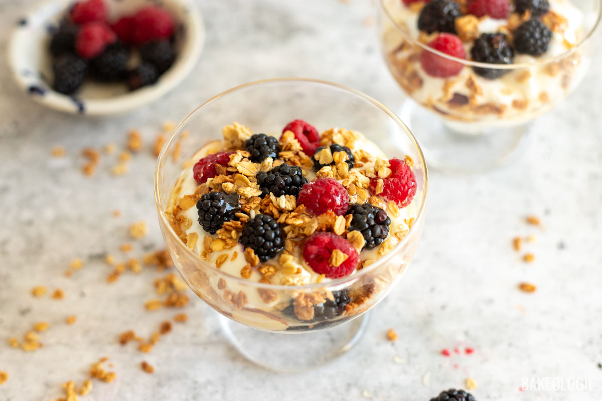 Side view of a clear glass filled with layers of creamy Greek yogurt, crunchy almond granola, fresh raspberries, and blackberries, garnished with extra berries and granola scattered around the glass.