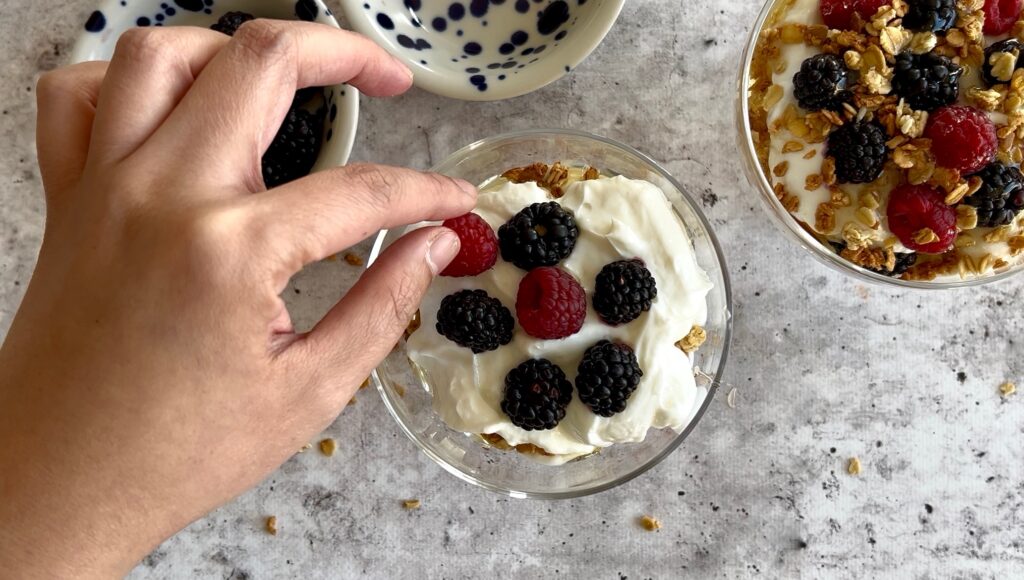 A hand placing fresh raspberries and blackberries onto a layer of creamy Greek yogurt in a glass, assembling a parfait.
