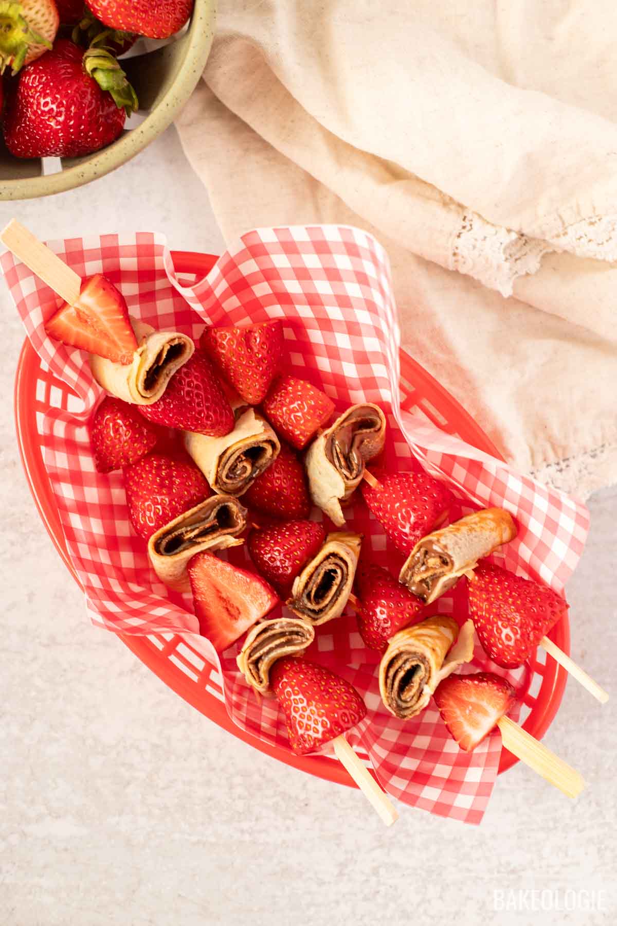 Overhead view of strawberry and hazelnut crepe skewers in a red basket lined with checkered paper, featuring rolled crepes with chocolate spread and fresh strawberries on wooden picks — a playful and easy summer dessert idea.