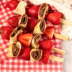 Close-up of strawberry and hazelnut crepe skewers arranged on red gingham parchment paper, featuring sliced rolled crepes with chocolate hazelnut spread and halved strawberries on wooden picks.